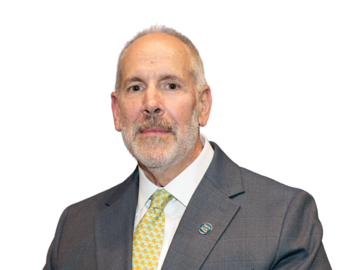 Older white male presenting with a short beard, in a grey suit and yellow tye